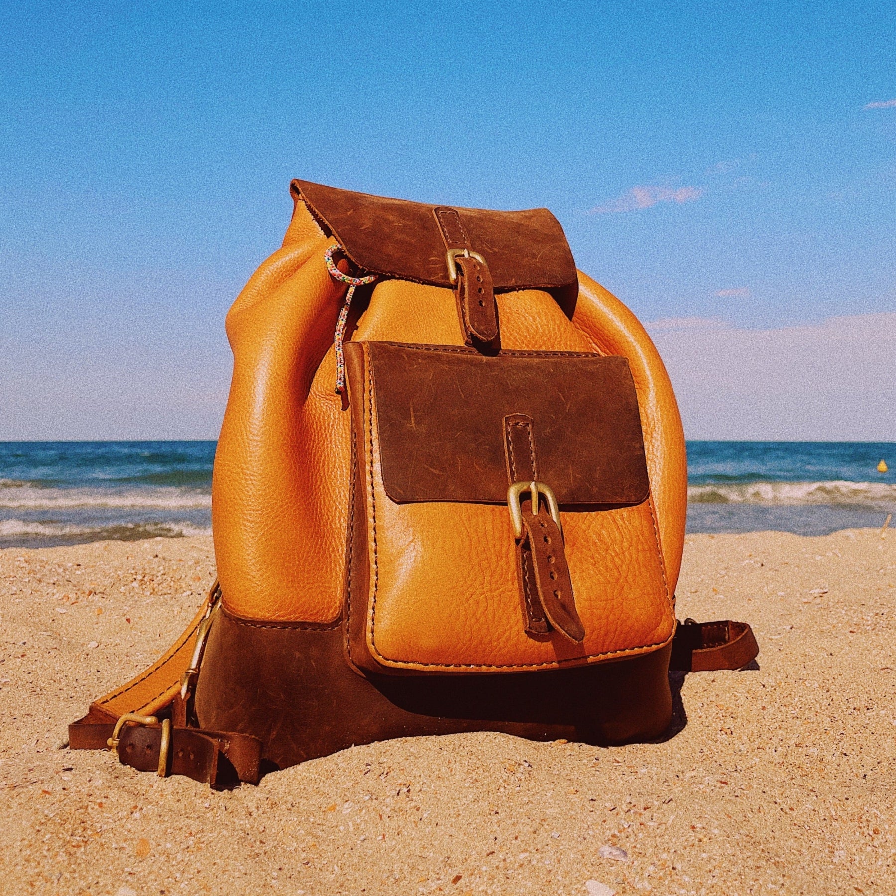 Brown leather backpack on a sandy beach with ocean in the background
