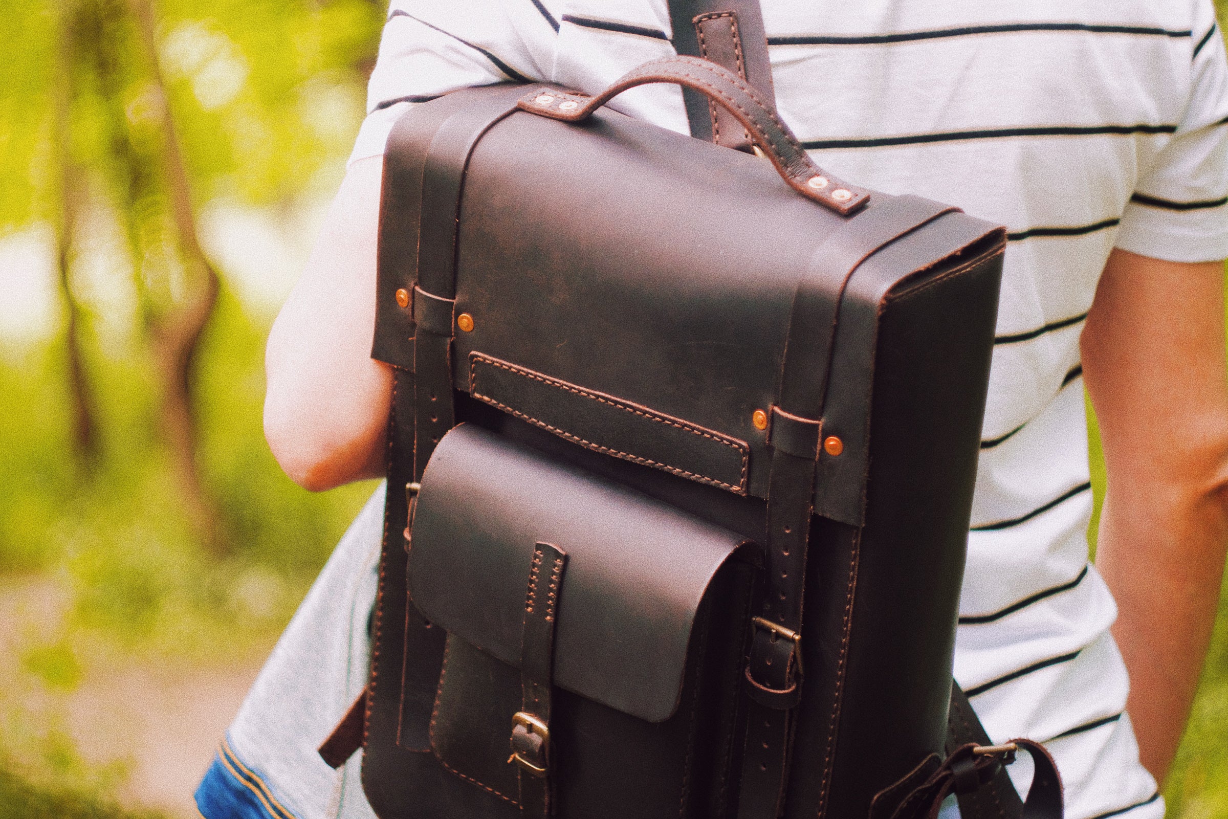 Person holding a brown leather backpack outdoors with greenery in the background