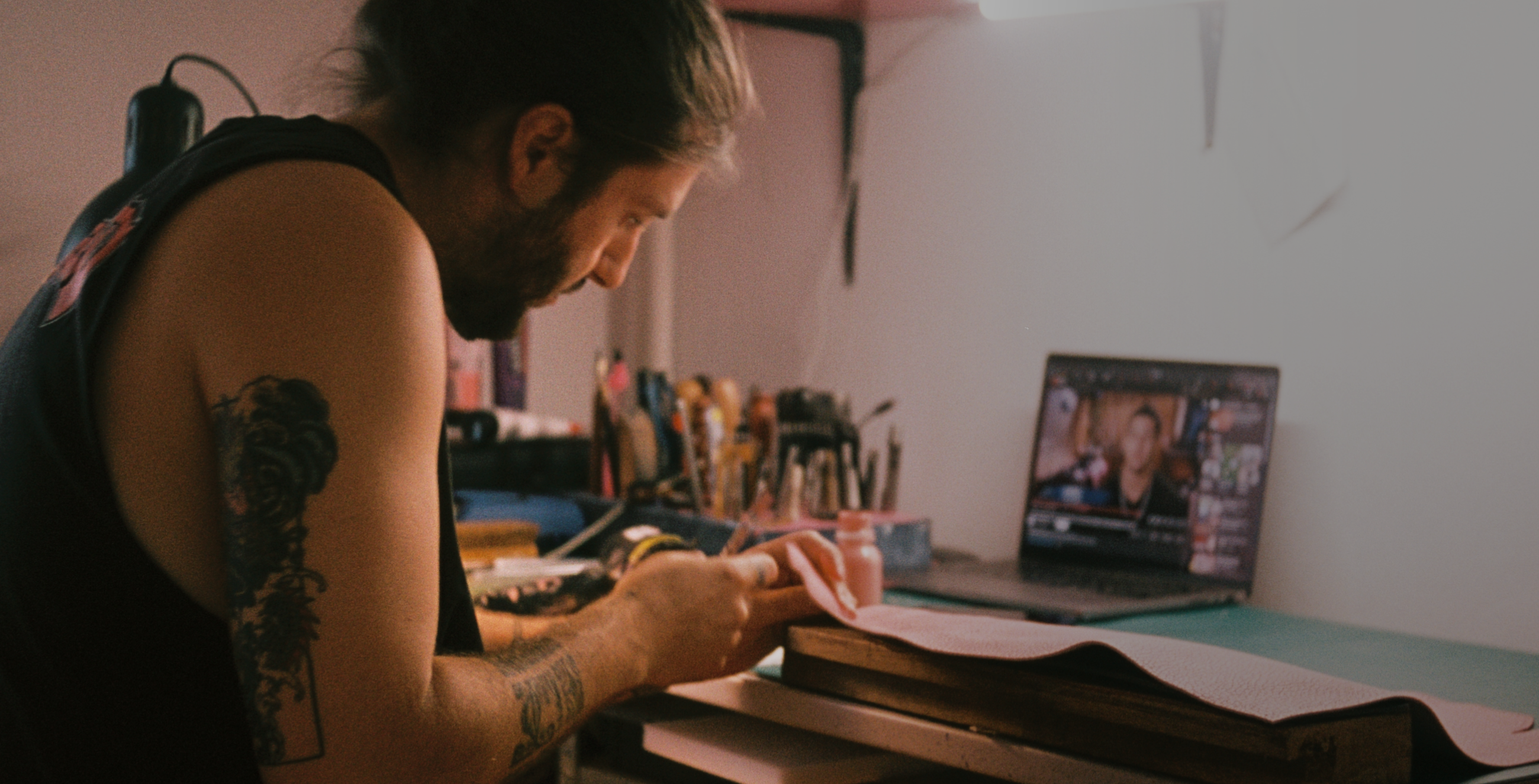 Man with tattoos working on a leather project by hand on a desk with leather tools.